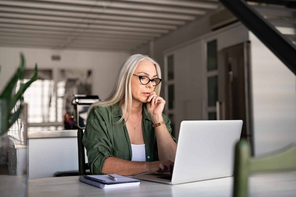 Focused old woman with white hair at home using laptop. Senior stylish entrepreneur with notebook and pen wearing eyeglasses working on computer at home. Serious woman analyzing and managing domestic bills and home finance.