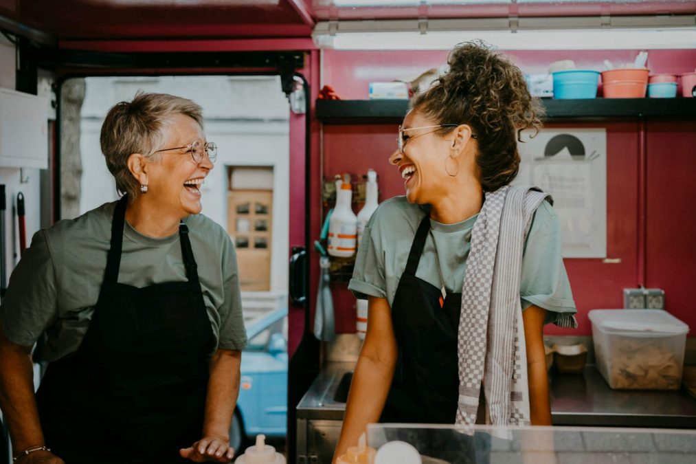Duas mulheres empreendedoras rindo apoiadas na bancada de atendimento de uma lanchonete - negócio e empreendedorismo